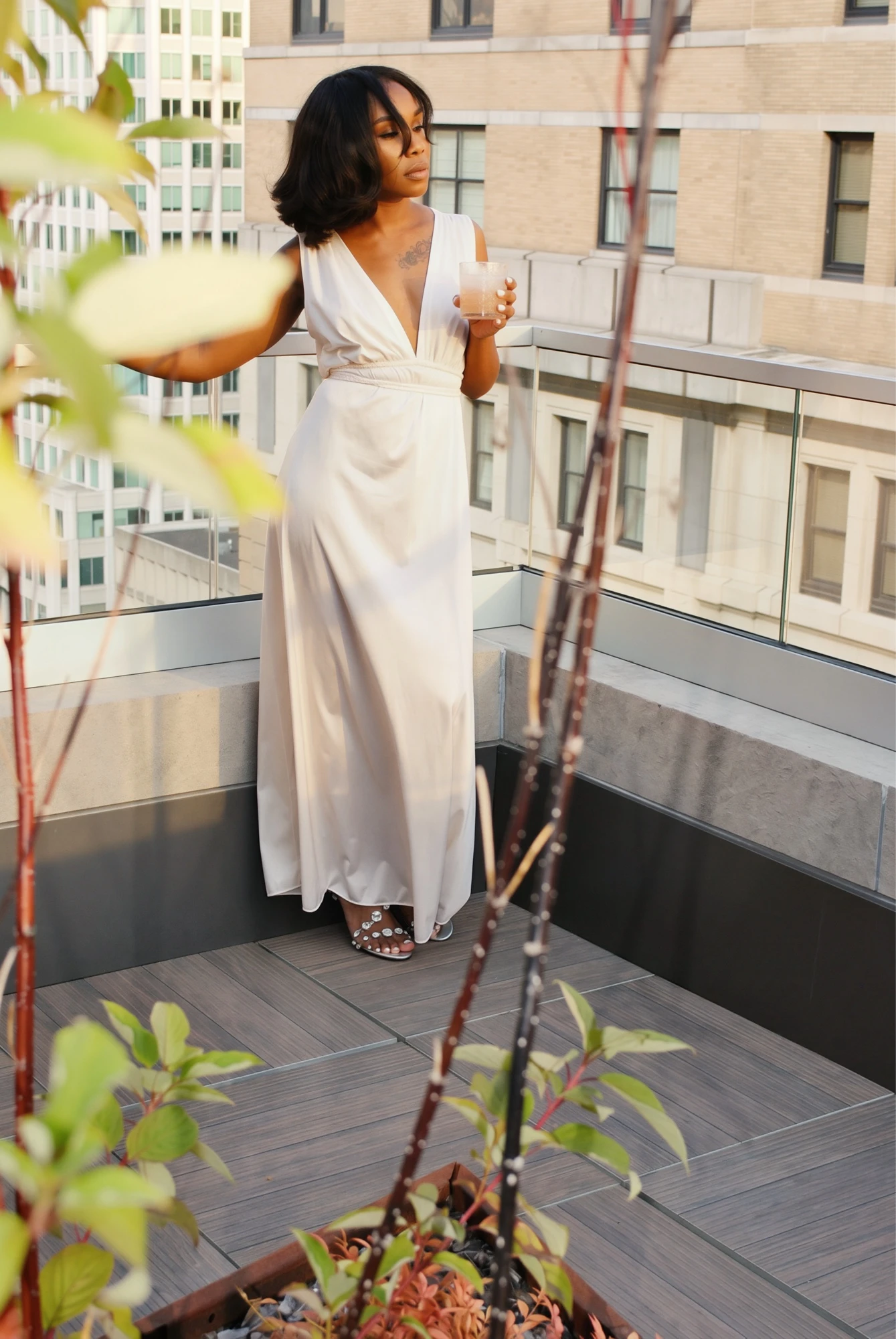 Full-length portrait in a flowing white deep-V gown and embellished sandals on a sunlit balcony framed by plants