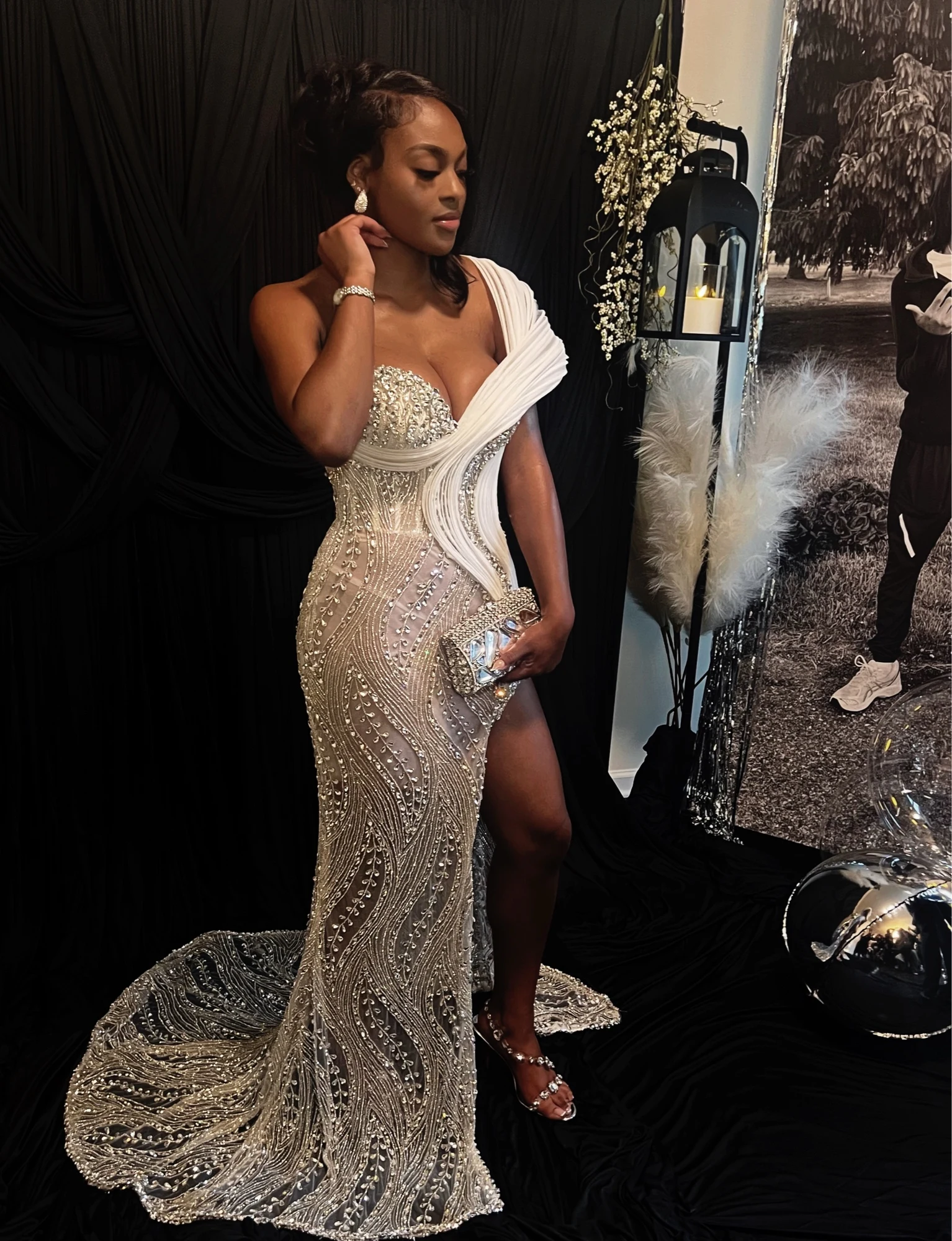 Crystal-beaded evening gown with sculptural white shoulder draping, thigh-high slit, and sweeping train against black curtains